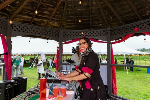 A DJ in a gazebo smiles while mixing music, surrounded by festive decorations and a lively outdoor event.