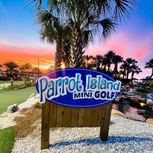 Sign for Parrot Island Mini Golf with palm trees and a colorful sunset in the background.