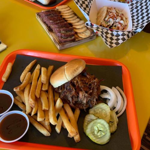A tray with a pulled pork sandwich, fries, pickles, onions, and two cups of barbecue sauce.