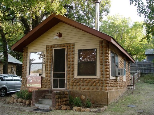 A small, rustic cabin with a stone exterior, surrounded by trees and a sign indicating it's available for rent.