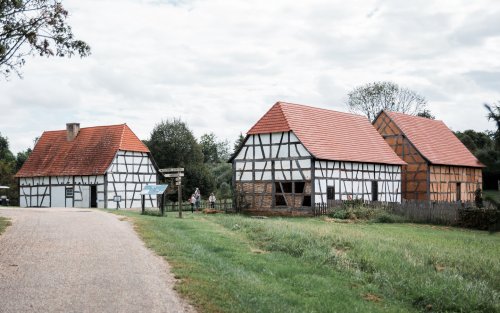 Three traditional timber-framed houses with red roofs beside a gravel road, surrounded by greenery and trees.