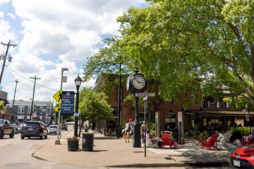 A sunny street scene featuring a clock, trees, and people enjoying outdoor seating near a brick building.