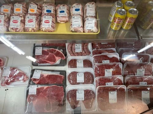 Display of various packaged meats, including steaks and ground beef, in a refrigerated case with cans in the background.