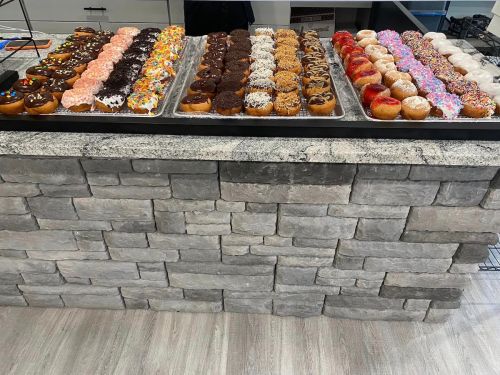 A display of colorful donuts arranged in rows on a countertop with a stone wall background.