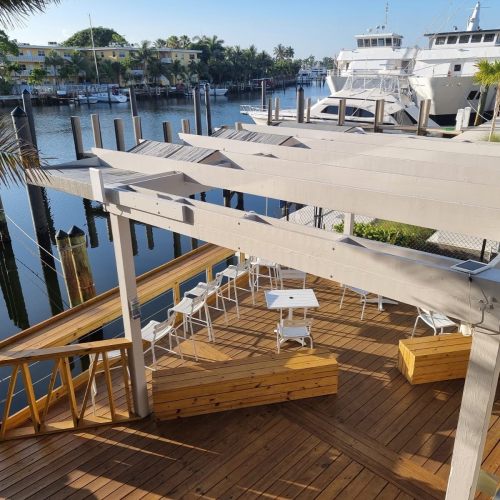 A wooden deck with white chairs overlooks a marina filled with boats and palm trees under a clear blue sky.