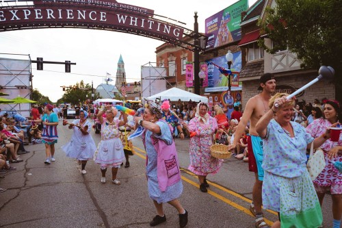 A lively parade scene with participants in colorful costumes, celebrating in a festive street under an archway.