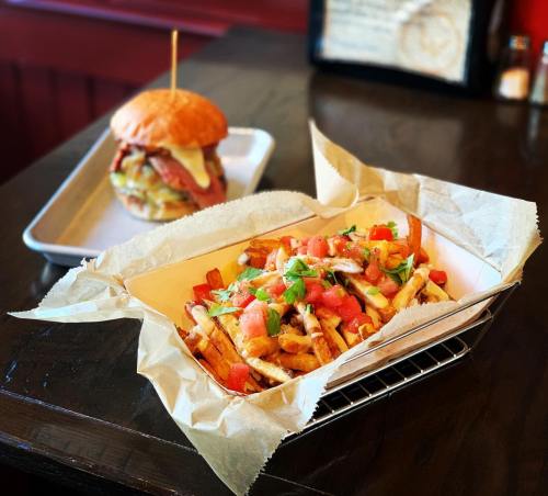 A basket of loaded fries topped with tomatoes and herbs, with a burger in the background on a wooden table.