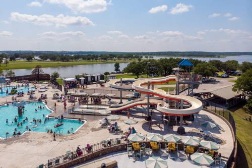 Aerial view of a water park with a large slide, swimming pool, and people enjoying the sunny day by a lake.