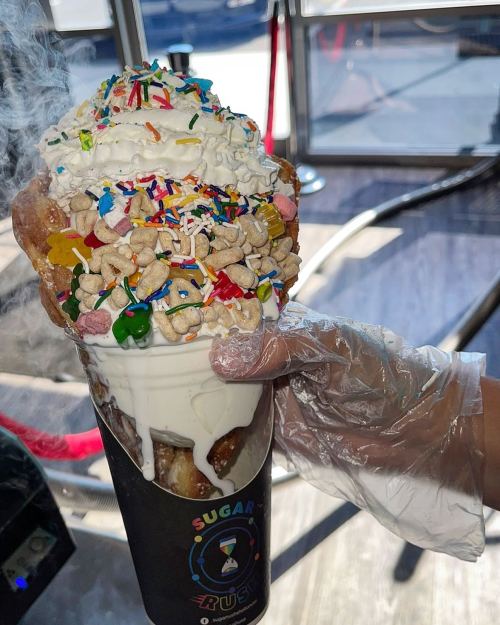 A hand holds a large ice cream cone topped with whipped cream, sprinkles, and colorful candies, with smoke rising.