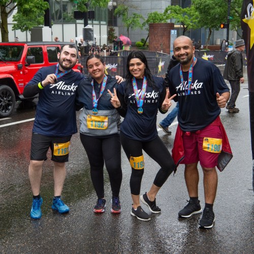 Four participants in matching Alaska Airlines shirts pose with smiles and peace signs at a rainy race event.