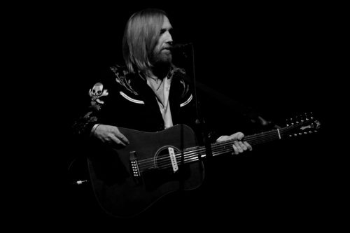 A musician with long hair performs on stage, holding an acoustic guitar and wearing a black jacket with skull designs.