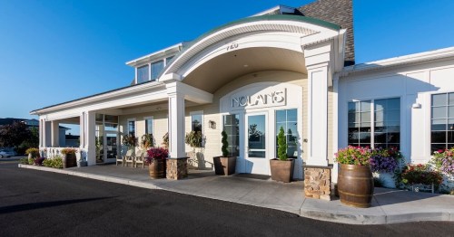Exterior view of a building with a welcoming entrance, surrounded by flowers and decorative barrels. Clear blue sky above.