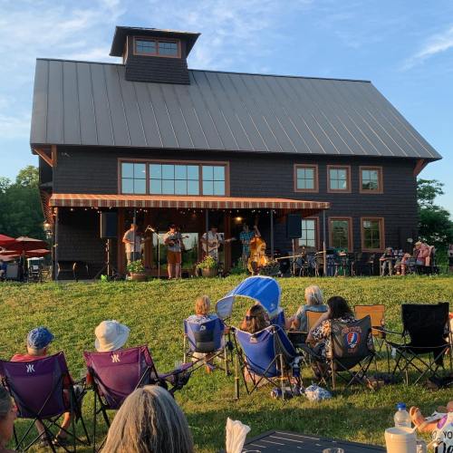 A live band performs on a stage in front of a large barn, with an audience seated on lawn chairs.