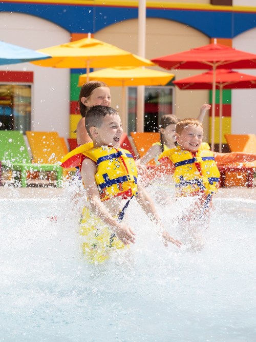 Children in life jackets joyfully splash in a pool, surrounded by colorful umbrellas and lounge chairs.