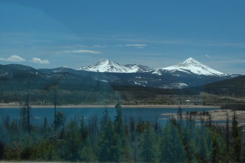 Snow-capped mountains rise above a serene lake, surrounded by lush green trees under a clear blue sky.
