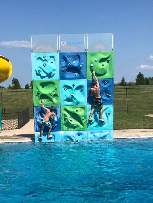 Two children climbing a colorful outdoor climbing wall beside a swimming pool on a sunny day.
