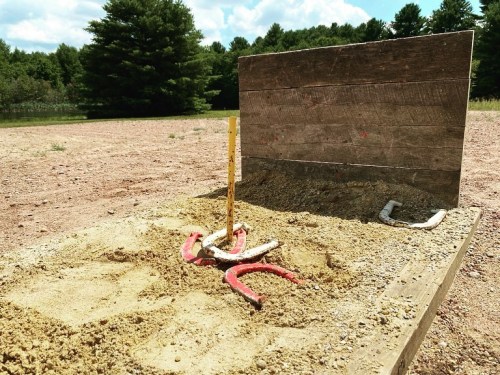 A sandy area with a wooden backboard and a horseshoe pit, featuring a yellow stake and red horseshoes.