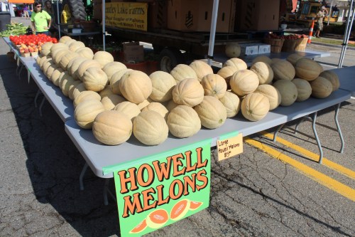 A table at a market displaying rows of melons with a sign reading "Howell Melons" and fresh produce in the background.