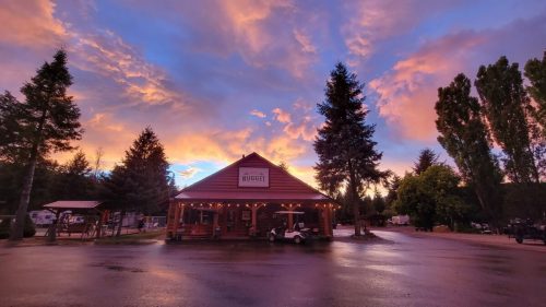 A rustic building with a sign reading "Nugget" under a vibrant sunset sky, surrounded by trees and a wet pavement.