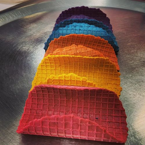 A stack of colorful waffle taco shells in vibrant shades of purple, blue, green, yellow, orange, and red on a silver tray.