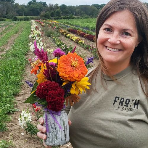 A smiling person holds a bouquet of colorful flowers in a field with rows of blooming plants in the background.