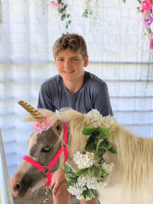 A young boy smiles next to a small unicorn adorned with flowers and a pink harness in a decorated setting.