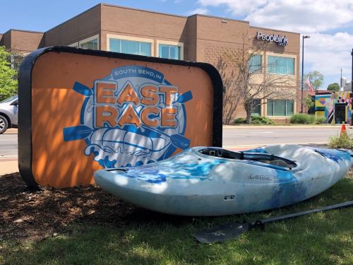 A kayak rests beside a large sign for the East Race in South Bend, IN, with a building in the background.