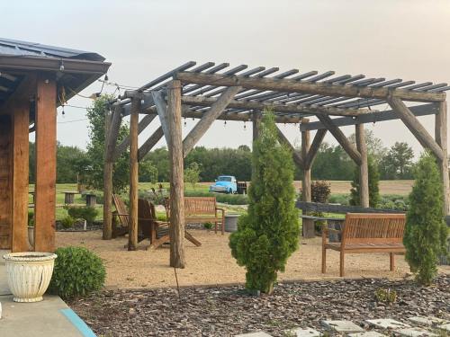 A wooden pergola with benches in a garden setting, surrounded by greenery and a distant blue car.