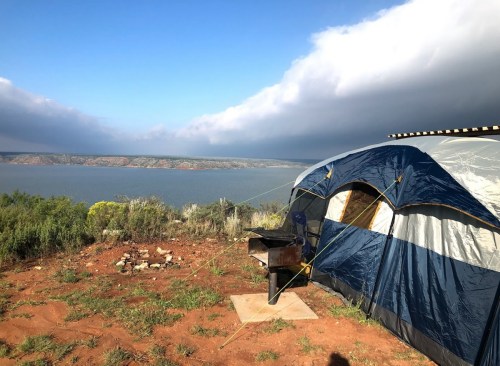 A tent set up near a lake, with a scenic view of the water and distant hills under a partly cloudy sky.