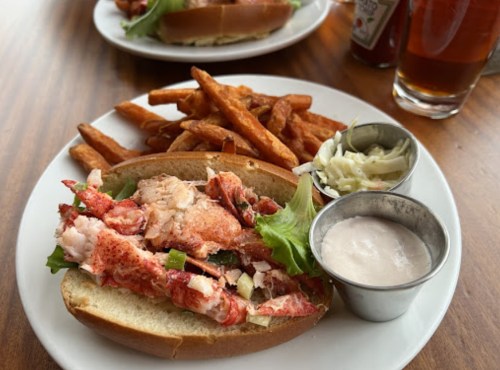 A lobster roll with lettuce, served with sweet potato fries, coleslaw, and a dipping sauce on a white plate.