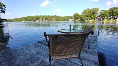 A peaceful lakeside view from a wooden dock with two chairs and a table, surrounded by trees and homes.