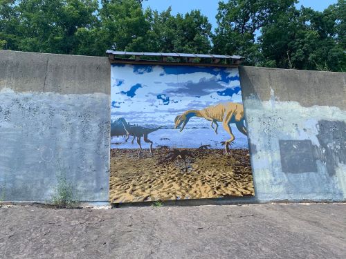 A mural depicting dinosaurs on a weathered concrete wall, surrounded by greenery and a blue sky.