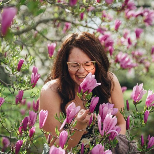A woman with glasses joyfully interacts with blooming pink magnolia flowers in a garden.
