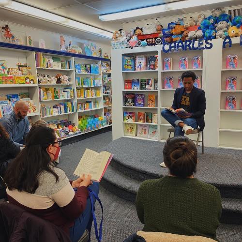 A man reads to an audience in a cozy bookstore, surrounded by colorful children's books and plush toys.