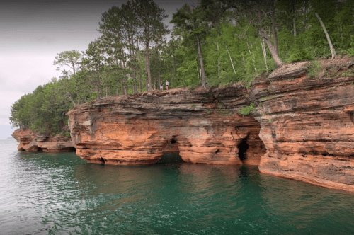 Rocky shoreline with green trees, featuring natural arches and calm turquoise water. Two people stand on the cliff.