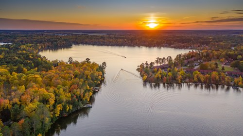 Aerial view of a serene lake at sunset, surrounded by colorful autumn trees and gentle boat trails.