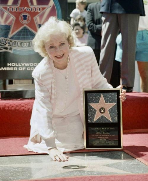 A smiling woman in a white outfit kneels beside a star on the Hollywood Walk of Fame, holding a plaque.