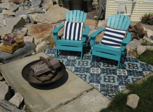 Two turquoise Adirondack chairs with striped cushions sit on a patterned mat near a fire pit surrounded by rocks.