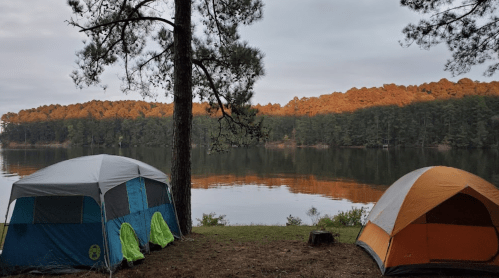 Two tents by a calm lake, surrounded by trees, with a soft morning light reflecting on the water.