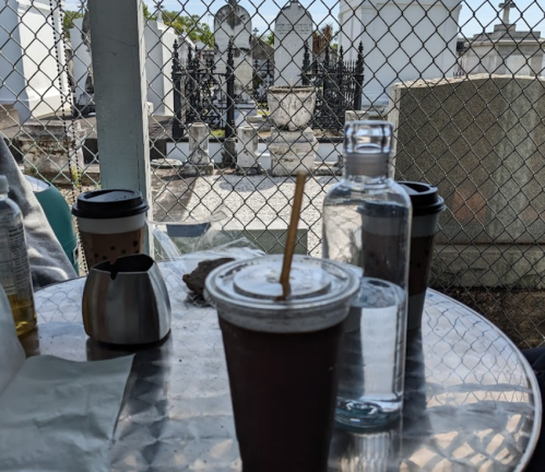 A table with drinks in a cemetery, featuring a coffee cup, a water bottle, and a view of gravestones in the background.