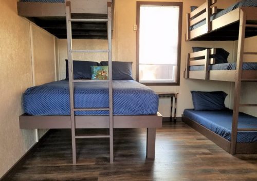 A cozy room with a double bed, bunk beds, and a small table, featuring blue bedding and wooden flooring.