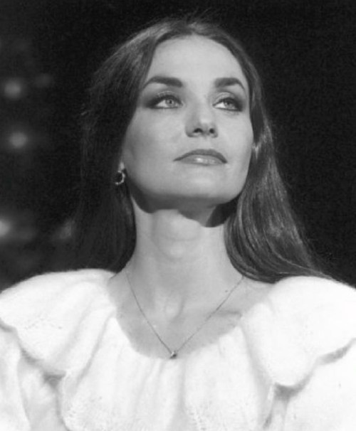 A woman with long hair and a white ruffled top gazes thoughtfully, captured in a black and white portrait.