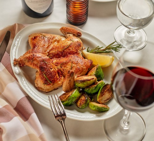 A plate of roasted chicken with lemon and Brussels sprouts, accompanied by a glass of red wine and elegant tableware.