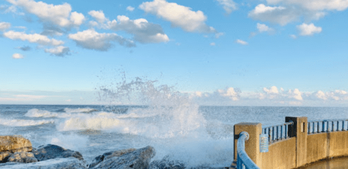 Waves crashing against a rocky shore under a blue sky with scattered clouds.