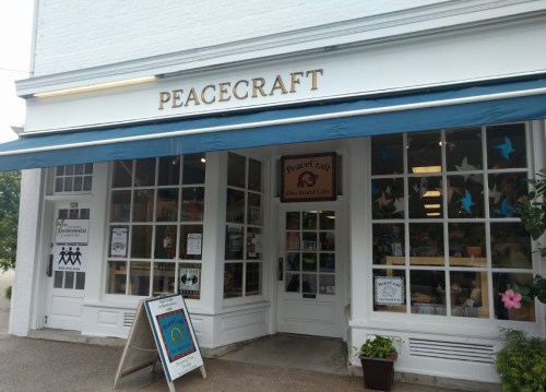 Exterior of PeaceCraft store with large windows, blue awning, and a sign indicating it's open. Colorful decorations visible.