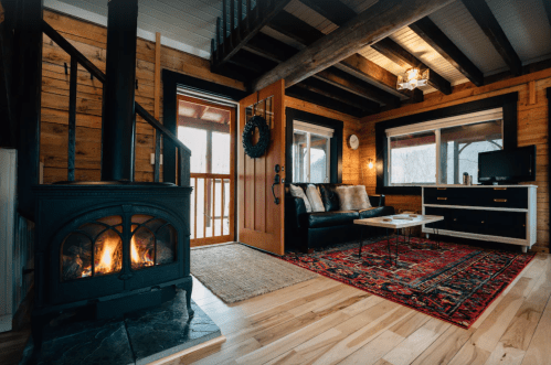 Cozy cabin living room with a wood stove, wooden beams, a rug, and large windows overlooking a scenic view.