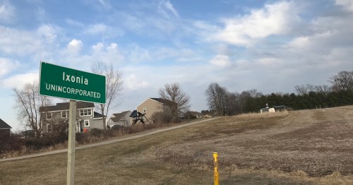 Sign for Ixonia, an unincorporated area, with houses and a cloudy sky in the background.