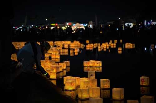 A person kneels by a pond, placing glowing lanterns on the water, surrounded by a crowd at night.