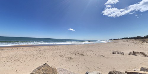 A sunny beach scene with gentle waves, a clear blue sky, and a few people in the distance along the shoreline.