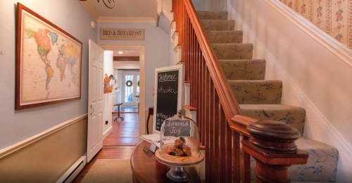 A cozy hallway with a staircase, a world map on the wall, and a chalkboard sign welcoming guests to a bed and breakfast.